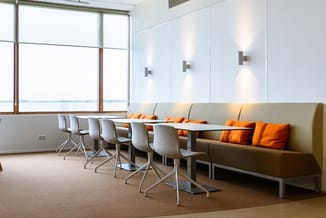 Modern kantoor met beige sofa met oranje kussens, vier grijze bureaustoelen aan kleine tafels, groot raam, witte muurpanelen