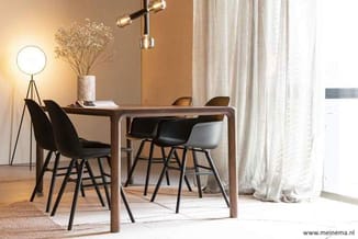Houten eettafel met zwarte stoelen, staande lamp, bloemen in vaas, gordijnen, houten vloer, warmwitte verlichting.