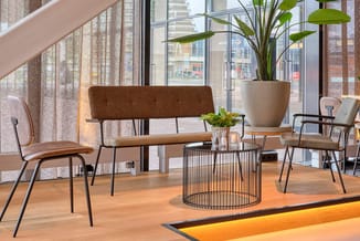 Modern lounge met bruine stoelen, houten bank, rond metalen tafeltje, grote plant in witte pot en ramen met gordijnen.