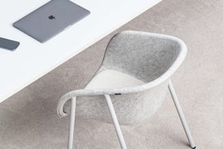 Grijze gestoffeerde stoel met witte poten op beige tapijt, met MacBook en smartphone op witte tafel op achtergrond.