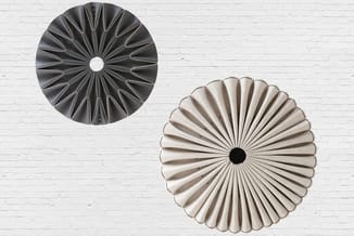 Twee decoratieve wandpanelen met radiale plissévouwen tegen witte bakstenen muur; één donkergrijs, één beige-wit.