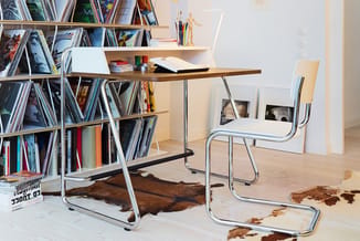 Werkplek met bruin houten bureau, zilveren metalen poten, witte bureelstoel, metalen boekenkast met magazines en boeken, hout