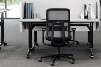 Zwarte mesh bureaustoel voor een kantoor met twee witte tafels op wielen tegen grijze muur met opberging en ramen.