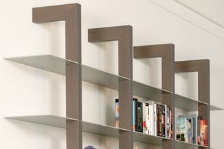 Boekenkast met houten frame in taupe/grijs en grijze schuine legplanken met boeken, tegen witte muur.
