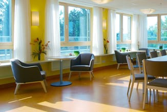 Modern kantoor met gele accenten, grote ramen, zwarte fauteuils, houten stoelen, lage tafel en boekenkasten langs de wand.