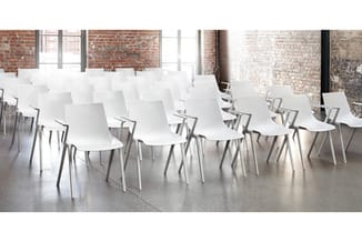Witte kunststof stoelen met aluminium poten in rijen opgesteld in een loft-achtige ruimte met bakstenen muren en betonvloer.