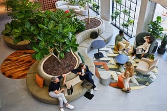 Modern kantoorinterieur met beige ronde zitbank rond plant, blauwe bijzettafels, losse stoelen en geomtrisch patroon vloerkle