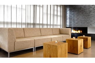 Beige loungebank met metalen frame, houten kubussen als bijzettafels, lineaire ramen en sfeerhaard op achtergrond.
