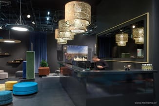 Modern receptie met zwarte balie, gouden filigree hanglampen, blauwe poef, groene plantenbak en wandmonitor boven balie.