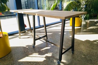Houten vergadertafel met zwart metalen onderstel en wieltjes in kantoorruimte met palmen en gele elementen.