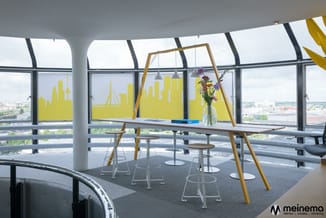 Modern kantoor op hoogte met gele skyline-ramen, houten vergadertafel, witte barkrukken, hangende verlichting en bloemenvaas.