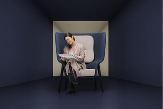 Vrouw zittend in blauwe/grijze fauteuil met hoge rug in minimalistische ruimte, notitieblok in handen.