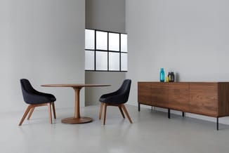 Minimalistisch interieur met ronde houten tafel, twee zwarte stoelen, houten dressoir en raam met zwart raamkozijn.