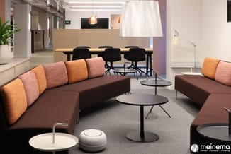 Modern kantoor met bruine modulaire bank met oranje kussens, ronde tafels, staande lamp, vergadertafel met zwarte stoelen en
