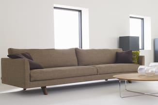 Grijsbruine lounge bank met houten poten in minimalistisch interieur met glazen deuren en houten tafel.