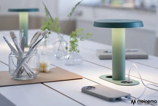 Moderne werkplek met groene tafellamp, glashouder met penselen, plant, smartphone en kantoormateriaal op wit bureau.