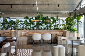 Moderne lounge met beige hoekbank, witte stoelen, ronde tafels en groene kantoorplanten tegen betonnen plafond.