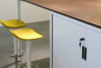 Twee gele barkrukken met witte poten bij een witte kastenwand met gesloten deuren en slot in kantooromgeving.