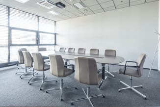 Moderne vergaderzaal met ovale tafel, grijze draaistoelen en grote ramen met lamellen. Grijze vloer, witte wanden.