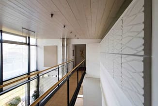 Moderne kantoorgang met houten plafond, raampartij, zwarte reling, houten handgreep en witte panelen aan rechterwand.