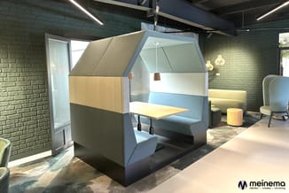 Akoestische belcel in blauwe en beige tinten met houten tafel, in modern kantoorlandschap met bakstenen wanden en hanglampen.