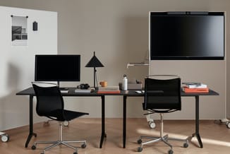 Moderne werkplek met zwart bureau, twee bureaustoelen, monitoren en driehoekige bureaulamp op houten vloer tegen beige wand.