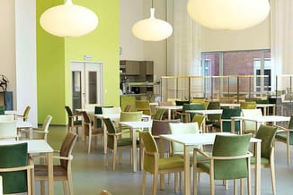 Moderne kantineruimte met houten stoelen in groen en taupe, witte ronde tafels, hangende design-lampen, groen accent-wand en