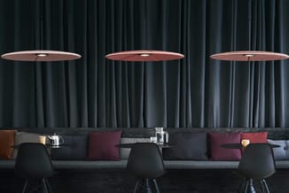 Drie ronde tafel met rode houten blad, zwarte stoelen en ophangende rode lamp boven grijze bank met paarse en bruine kussens