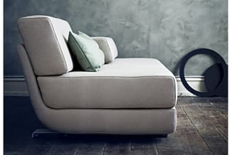 Grijsbeige loungezetel met afgeronde vormen, groene en beige kussens, op donkerhouten vloer tegen betonnen muur. Zwart cilind