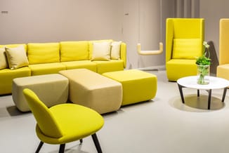 Showroom met gele zitmeubels: modulaire bank, loungestoel, poefs en witte ronde tafel met bloemen op neutrale ondergrond.