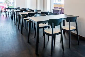 Moderne kantineruimte met zwarte houten tafels, zwarte stoelen met rieten zitting en lichte tafelbladen in rijen opgesteld.