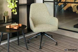 Grijsgroene loungestoel met textielupholstering op zwart onderstel, geplaatst op grijze tapijttegel naast zwart rond bijzetta