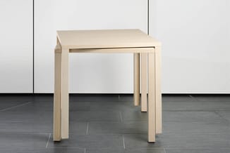 Lichte houten vierkante tafel met vier poten, staand op donkergrijze vloer tegen witte wand.