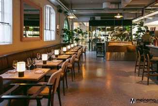 Modern restaurant met zwarte design-stoelen, houten tafels, zwarte zitbank, hangende verlichting en glazen wand naar bar.