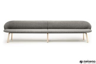 Grijze loungbank met afgerond design, gestoffeerd in gewatteerd textiel, op lichte houten poten. Meinema-logo onderin rechts.