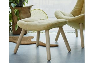 Crèmekleurige fauteuil en voetenbankje met houten poten in minimalistisch interieur met plant en kurk mat.