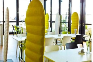 Moderne kantoorruimte met gele akoestische schermen tussen werkplekken, witte tafels, groene en zwarte stoelen, grote ramen.