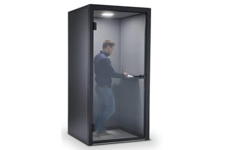 Man in donkerblauwe hoodie staat in grijze geluidsdichte belcel met glazen wand, zwart frame en LED-verlichting.