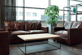 Bruine lederen bank en fauteuil met zilveren noppen rond salontafel met wit blad in moderne kantoorlounge met grote ramen.