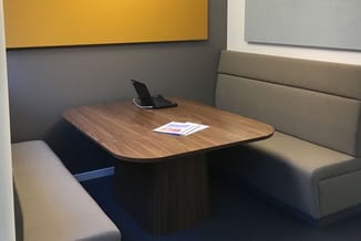 Hoekbank in beige stof met houten vergadertafel, laptopstandaard en documenten. Grijze wanden met gele en blauwe kleurklemmen