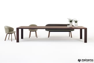 Donkerhouten vergadertafel met beige en grijze stoelen, decoratieve plantenpotten, en een beige fauteuil links.