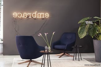 Moderne loungehoek met twee donkerblauwe fauteuils, houten voetenstukken, glazen bijzettafels, bloemenvaas en neonreclame "we