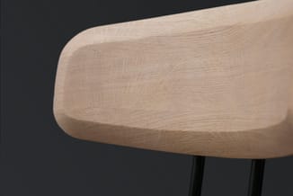 Houten stoel met afgeronde zitting in licht houtfinish, zwarte poten, detail van de zitting tegen donkere achtergrond.
