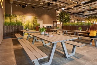 Moderne kantineruimte met houten picknicktafels en banken, groene plantenwanden, spotverlichting en houten vloer.