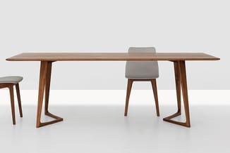 Houten vergadertafel met trapeziumvormige poten, grijze stoel en kruk in minimalistisch interieur.