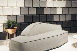 Grijze stoffen loungebank in moderne ruimte met zwart-wit blokkenpatroon wandbekleding en gouden plantenbak.