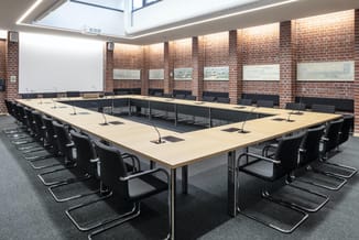 Rechthoekige vergadertafel met licht houten blad, zwarte stoelen, microfoons en schermen. Bakstenen wand, daglicht van bovena