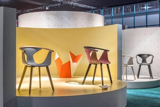 Tentoonstellingsstand met gele achterwand met moderne stoelen in zwart, rood en roze, houten poten, oranje sculptuur centraal
