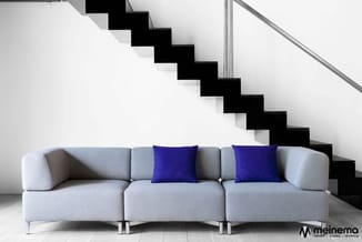 Grijze modulaire bank met drie zitplaatsen, twee blauwe kussens, metallic poten, in moderne witte ruimte met zwarte trap.