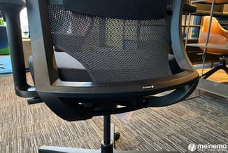 Zwarte ergonomische bureaustoel met mesh-rugsteun en zwart onderstel, geplaatst in een kantoor met grijze vloerbedekking en h